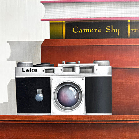 Detail of "Camera Shy" 2025 - a painting by James Carter at Nature's Studio - showing a Leica camera on a wooden surface with books stacked behind